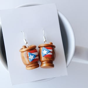 May include: A pair of dangle earrings featuring a miniature wooden barrel with the Puerto Rican flag painted on it. The earrings are silver and have a hook closure.