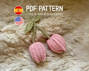 May include: A pair of pink crocheted tulip flowers with green stems and leaves. The image includes the text "PDF PATTERN + PRINTABLE STICKERS!" and flags of Spain and the United States.
