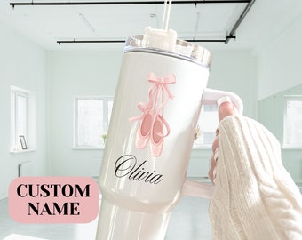 May include: White stainless steel tumbler with a pink ballet shoe design and the name "Olivia" printed on the side. The tumbler has a lid and a handle.