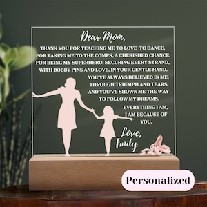 May include: A white acrylic plaque with a pink silhouette of a mother and daughter holding hands. The plaque has a message that reads "Dear Mom, Thank you for teaching me to love to dance, for taking me to the comps, a cherished chance. For being my superhero, securing every strand, with bobby pins and love, in your gentle hand. You've always believed in me, through triumph and tears, and you've shown me the way to follow my dreams. Everything I am, I am because of you. Love, Emily". The plaque is sitting on a wooden base.