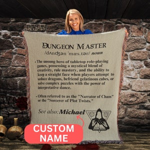 May include: A beige throw blanket with a definition of "Dungeon Master" printed on it. The definition includes the text "Narrator of Chaos" and "Sorcerer of Plot Twists". The blanket also has a custom name field and a black and white illustration of a hooded figure with a book and a pentagram.