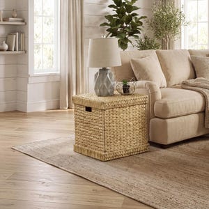 May include: A square storage ottoman crafted from woven light brown seagrass, featuring a flat lid and a cutout handle on the front side. The ottoman serves as a base for a gray geometric table lamp with a beige fabric drum shade.