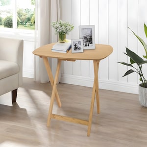 May include: A light-colored wooden folding table with a smooth, rounded rectangular top and cross-legged base. On the table sits a clear glass vase with white flowers, a stack of books, and two silver-framed photographs displaying black and white images.