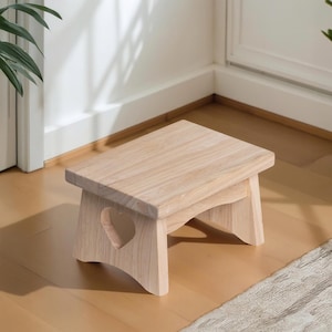 May include: A small, light-colored wooden step stool with a rectangular top and heart-shaped cutout on the side. The stool is made of natural wood and has a simple, minimalist design. It is suitable for children or adults.
