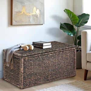 May include: A large, dark brown wicker storage trunk with a woven lid. The trunk is sitting on a light brown rug in a living room setting. There are books and headphones on top of the trunk.