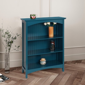 May include: A teal blue bookshelf with three shelves. The bookshelf has a slightly arched top and is made of wood. There are decorative items on the shelves, including a vase with white flowers, a glass jar, and a small potted plant.