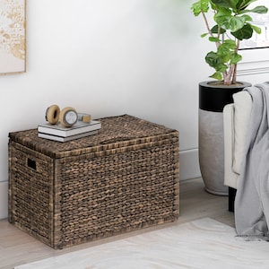 May include: A brown woven storage bin with a lid. The bin is sitting on a light wood floor with a rug in front of it. There are headphones and a book on top of the bin.