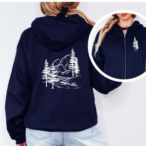 Puede incluir: Sudadera con capucha azul marino con cremallera y un gráfico blanco de montañas y árboles en la espalda. El pecho izquierdo presenta un diseño de árbol más pequeño. La sudadera tiene una capucha con cordón y una cremallera completa.