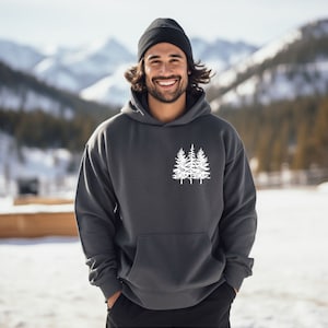 Peut inclure: Un sweat à capuche gris anthracite avec un motif blanc de trois sapins sur le devant. Le modèle porte un bonnet noir.