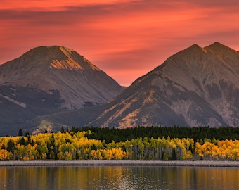 May include: A scenic landscape featuring majestic mountains under a vibrant, orange-red sky. The foreground showcases a forest with trees displaying autumn colors, reflected in the calm water below. A serene nature scene.