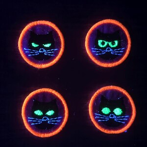 May include: Four circular embroidered patches featuring a black cat silhouette with glowing green eyes and white whiskers. Each patch is framed by a vibrant orange stitched border, creating a high-contrast design against the dark background.