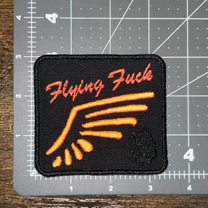 May include: Black square patch with orange text "Flying Fuck" and an orange wing design. A small skull is in the lower right corner. The patch is approximately 2.5 inches wide and 2.5 inches tall.
