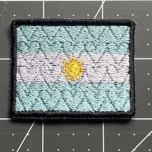 May include: Embroidered patch featuring the flag of Argentina. The rectangular patch has a light blue and white striped design with a yellow sun in the center. The edges are bordered with black stitching.