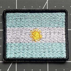 Puede incluir: Parche bordado con la bandera argentina. El parche rectangular tiene franjas celeste en la parte superior e inferior, una franja central blanca y un emblema de sol amarillo. El parche está bordeado con costuras negras.