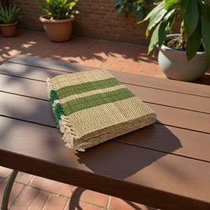 Puede incluir: Un textil rectangular plegado, tejido con rayas verdes y un patrón de espiga beige. Los bordes tienen flecos. El textil está sobre una superficie marrón.