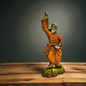 Handmade clay figurine of a Baul folk musician in an orange robe, holding a lantern and a small drum, displayed on a rustic wooden surface with a dark gradient background.