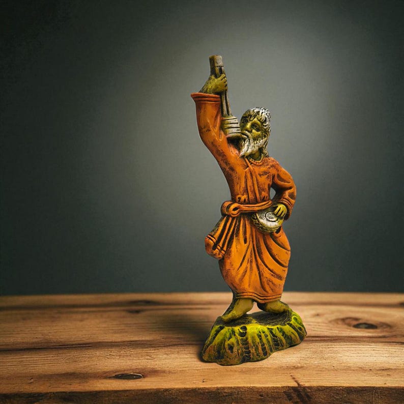 Handmade clay figurine of a Baul folk musician in an orange robe, holding a lantern and a small drum, displayed on a rustic wooden surface with a dark gradient background.