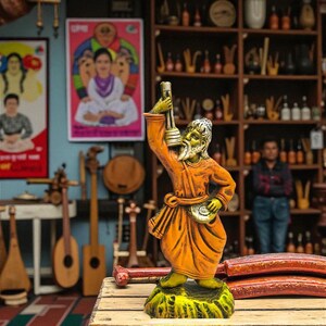 Handcrafted clay figurine of a Baul musician holding a lantern, painted in bright orange and yellow, displayed on a wooden table with traditional musical instruments and colorful posters in the background.