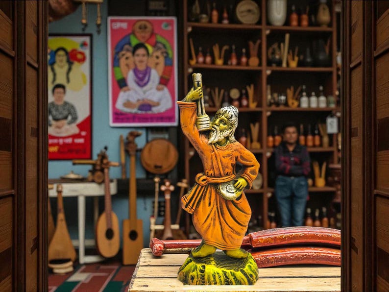 Handcrafted clay figurine of a Baul musician holding a lantern, painted in bright orange and yellow, displayed on a wooden table with traditional musical instruments and colorful posters in the background.
