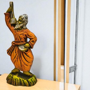 Terracotta Baul folk musician statue in orange attire holding an ektara, placed on a light wooden surface with a mirror and colorful vertical textile threads in the background.
