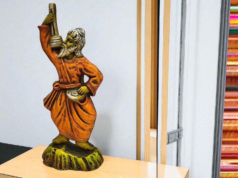 Terracotta Baul folk musician statue in orange attire holding an ektara, placed on a light wooden surface with a mirror and colorful vertical textile threads in the background.