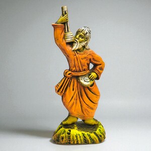 Handcrafted clay figurine of a Baul folk musician in an orange robe, holding a lantern and a small hand drum, standing on a textured green base against a light grey background.