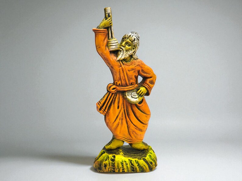Handcrafted clay figurine of a Baul folk musician in an orange robe, holding a lantern and a small hand drum, standing on a textured green base against a light grey background.