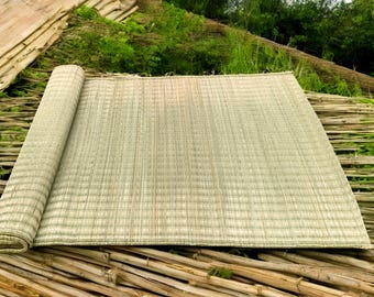 May include: A rolled-up, rectangular woven mat made of natural fibers, with a light beige color. The mat is partially unrolled, revealing its textured surface. The background features bamboo and greenery, suggesting an outdoor setting.