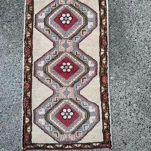 May include: Rectangular rug with a cream-colored background and a repeating geometric pattern. The pattern features diamond shapes in red, gray, and pink, with floral-like details. The rug has a brown border and fringe.