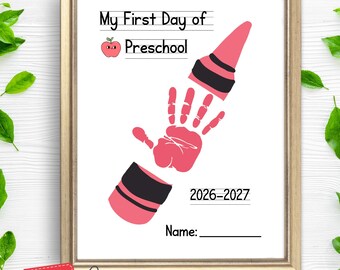 May include: A framed print featuring the text My First Day of Preschool, a small red apple icon, and a pink handprint graphic positioned between two segments of a pink and black crayon. The print includes the years 2026-2027 and a line for a name.
