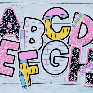 May include: A collection of colorful alphabet stickers, each designed with a unique pattern and school-themed illustrations. The letters A, B, C, D, E, F, G, and H are visible, featuring elements like pencils, notebooks, and a sneaker. The stickers are set against a light blue wooden background.
