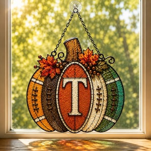 May include: A stained glass pumpkin suncatcher featuring a central orange panel with a white letter T, flanked by textured panels in yellow, brown, white, and green. The design includes football-style laces and orange maple leaf accents.