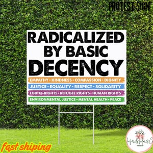 Pode incluir: Uma placa de jardim branca com o texto "RADICALIZED BY BASIC DECENCY" em preto. Abaixo, linhas coloridas listam valores como "EMPATHY", "JUSTICE" e "LGBTQ+ RIGHTS". A placa é exibida em um suporte de metal em frente a uma sebe verde.