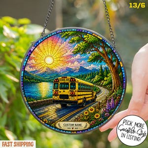 May include: A circular suncatcher featuring a yellow school bus driving on a road near a lake, mountains, and trees under a bright sun. The design includes a banner at the bottom with the text CUSTOM NAME and a blue decorative border frame.