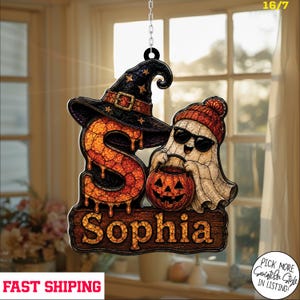 May include: A hanging suncatcher ornament featuring a large orange letter S topped with a black witch hat, next to a white ghost wearing a red beanie and sunglasses while holding a jack-o'-lantern. A wooden sign below reads Sophia.