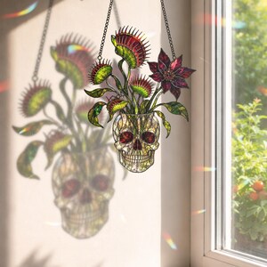 May include: A stained glass suncatcher featuring a human skull base holding several green and red Venus flytrap plants and a dark purple flower. The piece is suspended by a black metal chain and casts a colourful shadow on the wall nearby.