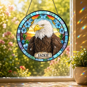 May include: A circular stained glass suncatcher featuring a detailed bald eagle with a yellow beak and white head feathers. The eagle is framed by blue flowers and a border of blue glass segments. A banner at the bottom displays the name JACKIE.