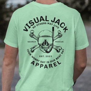 May include: A light green t-shirt featuring a black graphic of a skull wearing a scuba mask and crossed bones. The text "VISUAL JACK" is above the skull, and the text "SO OTHERS MAY LIVE" is below the skull. The text "SEARCH" is on the left side of the skull, and the text "RESCUE" is on the right side of the skull. The text "EST. 2023" is below the skull. The text "YOUR WORST DAY IS OUR BEST DAY" is in a circle around the skull. The text "APPAREL" is below the circle.