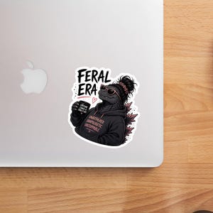 Op de afbeelding: Een sticker met een Godzilla-achtig wezen met een zonnebril en een zwarte hoodie. De hoodie heeft de tekst "UNBOTHERED UNAPOLOGETIC UNSTOPPABLE". De sticker heeft ook de woorden "FERAL ERA" en een koffiekopje met de tekst "CAFFEINE, CHAOS & ZERO FUCKS".