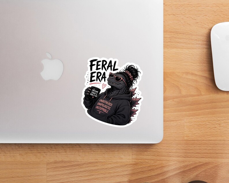Op de afbeelding: Een sticker met een Godzilla-achtig wezen met een zonnebril en een zwarte hoodie. De hoodie heeft de tekst "UNBOTHERED UNAPOLOGETIC UNSTOPPABLE". De sticker heeft ook de woorden "FERAL ERA" en een koffiekopje met de tekst "CAFFEINE, CHAOS & ZERO FUCKS".