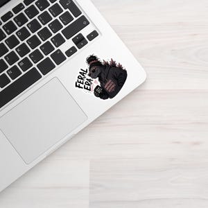 Op de afbeelding: Een sticker met een gestileerde illustratie van een gorilla met een zonnebril en een zwarte hoodie. De sticker heeft de woorden "FERAL ERA" in een vetgedrukte, zwarte letter. De gorilla houdt een klein object met tekst vast. De sticker staat op een laptop.