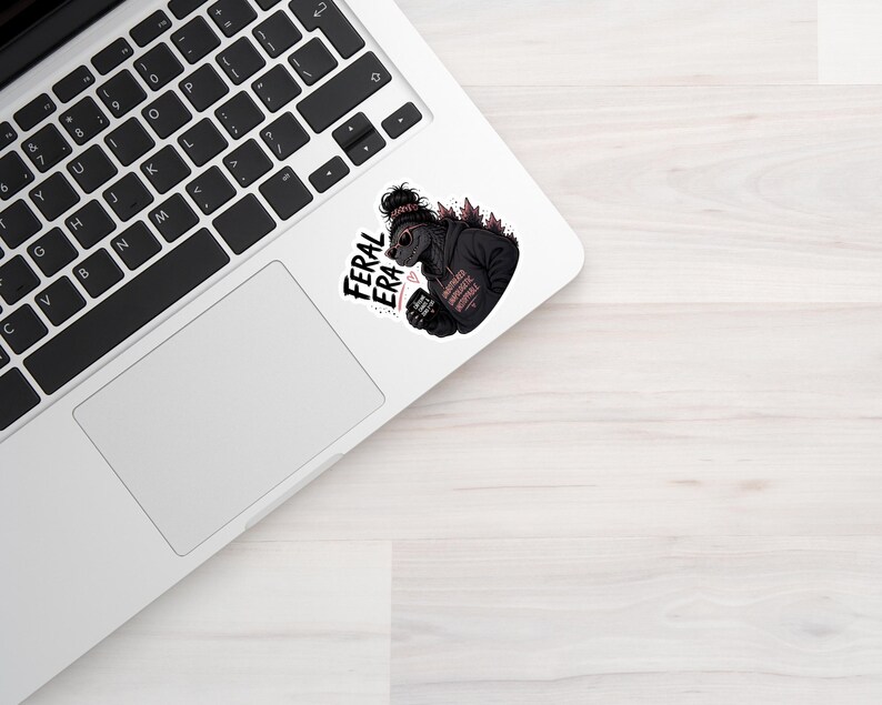 Op de afbeelding: Een sticker met een gestileerde illustratie van een gorilla met een zonnebril en een zwarte hoodie. De sticker heeft de woorden "FERAL ERA" in een vetgedrukte, zwarte letter. De gorilla houdt een klein object met tekst vast. De sticker staat op een laptop.
