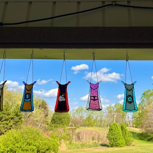 May include: Seven stained glass superhero cape ornaments hanging from a porch. Each cape features a unique design and color scheme, including yellow, blue, red, pink, teal, and green. The capes are suspended by chains against a backdrop of a blue sky and trees.