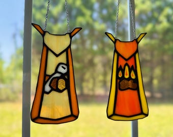 May include: Two stained glass suncatchers shaped like capes with black lead lines. The left cape is pale yellow and orange with a white and brown axe icon. The right cape is bright orange and yellow with a brown and yellow paw print icon.