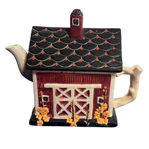 Puede incluir: Una tetera de cerámica con forma de granero rojo, techo de tejas negras, chimenea y puertas dobles blancas. La estructura presenta detalles florales amarillos en la base, un pico vertedor claro con forma de rama y un asa a juego.