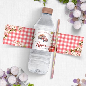 Op de afbeelding: Een waterfleslabel met rood-wit geruit patroon met een rode vrachtwagen die appels vervoert en de tekst "Cassie's Baby Shower The Apple of Our Eye".