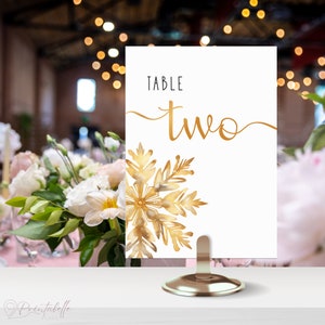 Table Number in small gold holder on a table. The design of the table number is elegant minimalistic. It is very clean and neat. There is nothing else on the sign except for the large snowflake and the words table two.