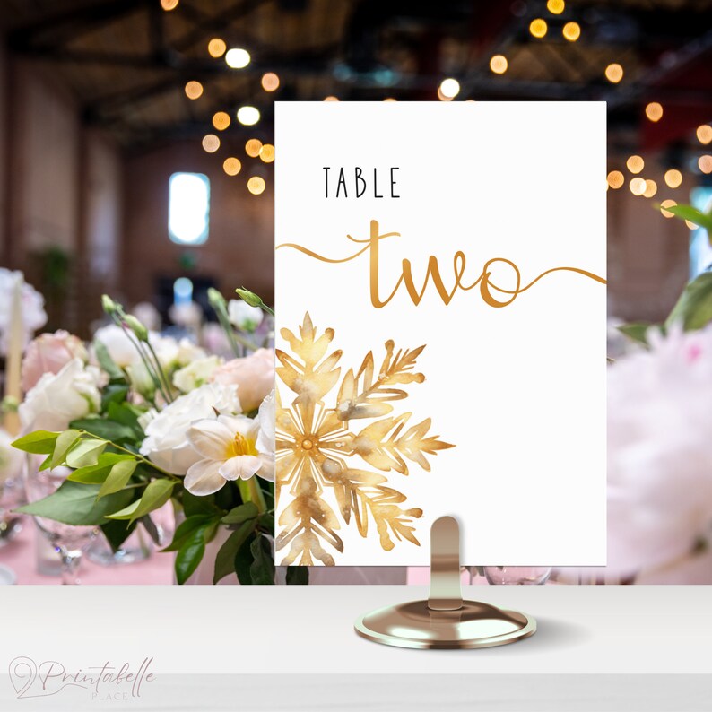 Table Number in small gold holder on a table. The design of the table number is elegant minimalistic. It is very clean and neat. There is nothing else on the sign except for the large snowflake and the words table two.