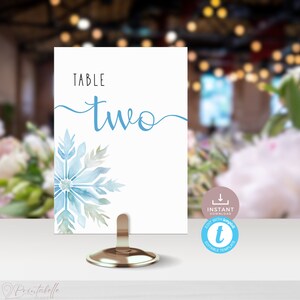 May include: Table number two with a blue and green watercolor snowflake design. The text "TABLE two" is written in a cursive font. The card is on a gold metal stand.  The text "INSTANT DOWNLOAD EDIT WITH TEMPLATES EDITABLE TEMPLATE" is on the card.