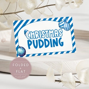 Może przedstawiać: Złożona karta "Christmas Pudding" z niebiesko-białymi ukośnymi paskami i świątecznymi ozdobami. Słowa "CHRISTMAS PUDDING" są pogrubione na niebiesko. Mała, okrągła, różowa naklejka z napisem "FOLDED + FLAT included" znajduje się w lewym dolnym rogu.